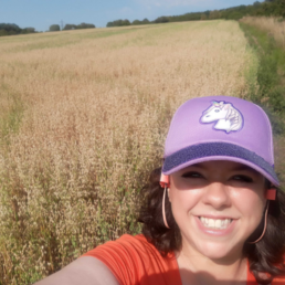 Danielle is wearing a cap with a unicorn on, standing in a field in the sunshine and grinning at the camera.